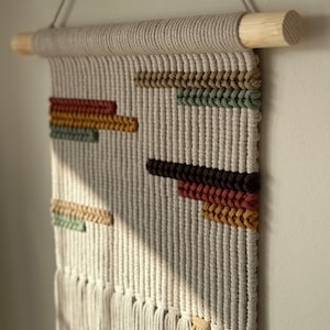 May include: A beige macrame wall hanging with a wooden dowel at the top. The macrame features geometric shapes in shades of brown, orange, yellow, and green.