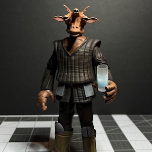 Puede incluir: Una figura de acción marrón y tostada de un personaje alienígena ficticio con cuatro ojos, sosteniendo un vaso transparente con líquido azul. La figura lleva un atuendo marrón y negro con un cinturón y botas marrones. Se ven tres manos adicionales en primer plano.