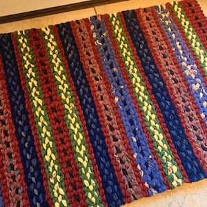 May include: A rectangular, handmade rug with vertical stripes in various colors. The rug features alternating stripes of red, blue, green, and yellow. The rug is made of woven fabric and has a textured appearance. The rug is placed on a tiled floor.