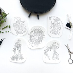 May include: Six black and white line drawings on white paper, each depicting a different skeleton figure. The figures include a skeleton holding a cat, a skeleton with flowers, a skeleton with a scythe, and three other skeletons in various poses.