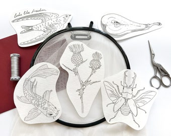Può includere: Un telaio da ricamo nero con tessuto a rete bianco, circondato da cinque ritagli di carta con disegni a inchiostro nero di un pesce koi, un cardo, uno scarabeo, un uccello con il testo looks like freedom e una pera, più filo d'argento e forbici.