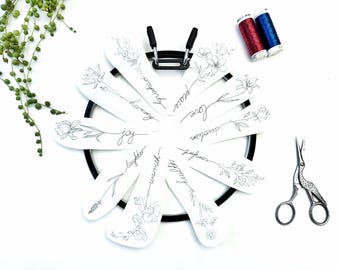May include: A black embroidery hoop holds ten white paper petals arranged in a circle. Each petal features a black line drawing of a flower with a cursive word: harmony, joy, laughter, warmth, health, comfort, devotion, love, peace, and gratitude.