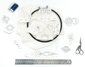 Könnte beinhalten: Ein schwarzer Stickrahmen mit weißem Stoff, umgeben von Papierschnitten mit Himmelsmotiven wie Sternen, Monden und einem Kometen. Der Text "LOOKING AT STARS?" ist sichtbar. Enthält zwei Garnrollen, eine Metallschere und ein 30 cm langes Lineal.