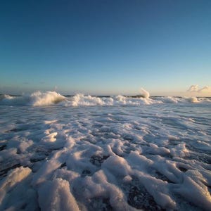 Può includere: Una vista dal basso di onde oceaniche che si infrangono su una riva sabbiosa, con schiuma di mare bianca che copre il primo piano. L'acqua passa dal blu profondo a tonalità più chiare vicino all'orizzonte sotto un cielo azzurro al tramonto.