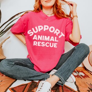May include: A bright pink t-shirt with the words "SUPPORT ANIMAL RESCUE" in white block letters. The shirt is worn with blue jeans and white sneakers. The person is sitting on a patterned blanket.