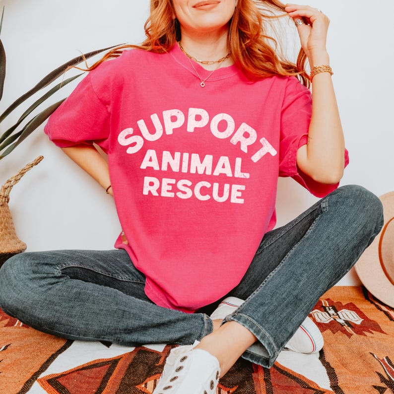 May include: A bright pink t-shirt with the words "SUPPORT ANIMAL RESCUE" in white block letters. The shirt is worn with blue jeans and white sneakers. The person is sitting on a patterned blanket.