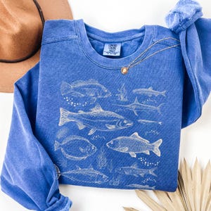 May include: A blue sweatshirt featuring a detailed white illustration of various fish species. The design includes different types of fish, seaweed, and bubbles. The sweatshirt has a crew neck and long sleeves. A gold necklace with a pendant is draped across the front.
