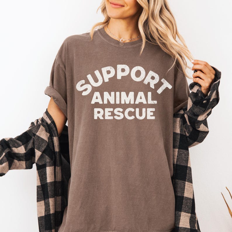 May include: A brown t-shirt with the words "SUPPORT ANIMAL RESCUE" in white block letters. The shirt is worn with a black and tan plaid flannel shirt. The sleeves of the t-shirt are rolled up.
