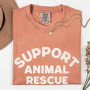 May include: A coral-colored t-shirt with the words "SUPPORT ANIMAL RESCUE" in large white letters. A gold star pendant necklace and a tan hat are also visible. The shirt has a Comfort Colors tag, size XL-TG-EG.