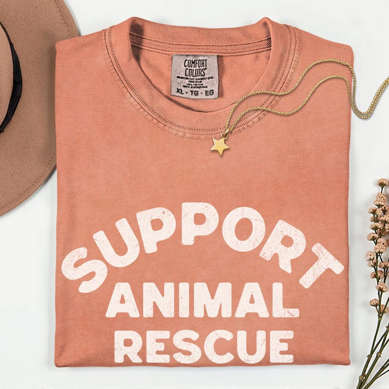 May include: A coral-colored t-shirt with the words "SUPPORT ANIMAL RESCUE" in large white letters. A gold star pendant necklace and a tan hat are also visible. The shirt has a Comfort Colors tag, size XL-TG-EG.