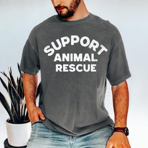 May include: A gray t-shirt with the words "SUPPORT ANIMAL RESCUE" in white block letters. The shirt is a solid color and has short sleeves. The person is wearing blue jeans and a watch.