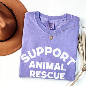 May include: A lilac-colored t-shirt with the words "SUPPORT ANIMAL RESCUE" in white block letters. A gold necklace with a pendant and a brown hat are also in the image. The shirt is folded and has a Comfort Colors tag.