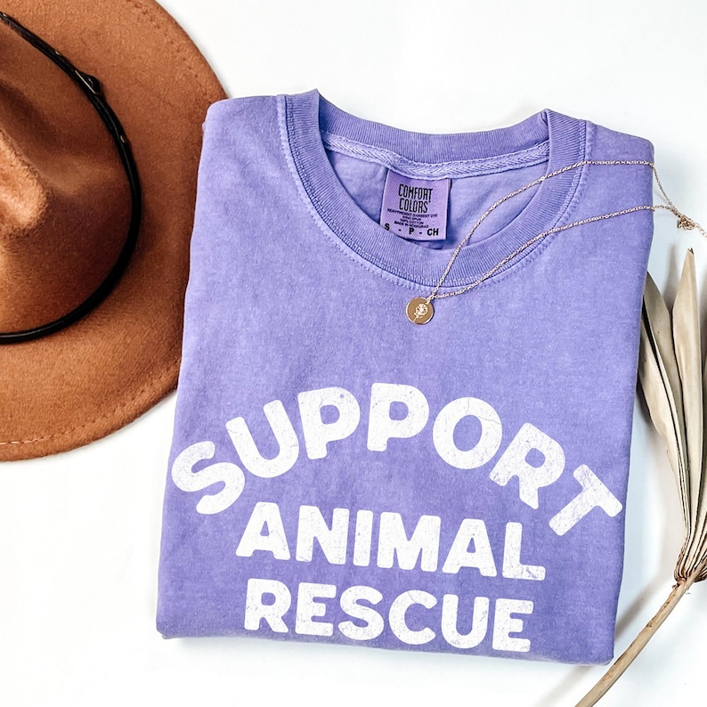 May include: A lilac-colored t-shirt with the words "SUPPORT ANIMAL RESCUE" in white block letters. A gold necklace with a pendant and a brown hat are also in the image. The shirt is folded and has a Comfort Colors tag.