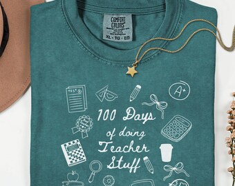 May include: Teal Comfort Colors t-shirt with white graphic text and illustrations. The shirt reads "100 Days of doing Teacher Stuff" with school-related images. A gold star pendant necklace is draped over the shirt.
