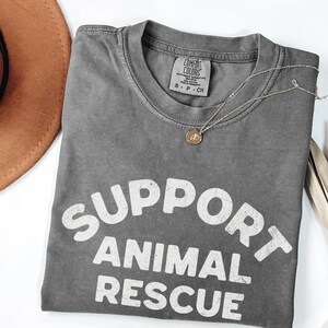 May include: A gray Comfort Colors t-shirt with the words "SUPPORT ANIMAL RESCUE" in white block letters. A silver necklace with a small pendant rests on the shirt. A brown hat is partially visible in the upper left corner.