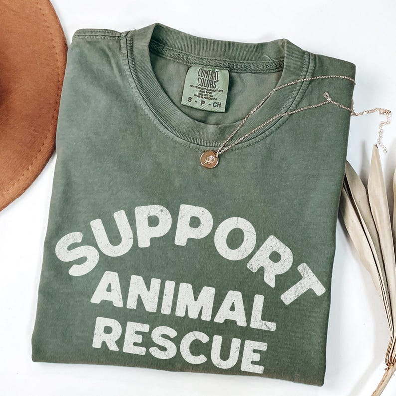 May include: An olive green t-shirt with the words "SUPPORT ANIMAL RESCUE" in white block letters. A gold necklace with a small pendant rests on the shirt. The shirt is folded and placed on a white surface.