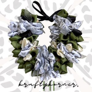 May include: A circular wreath made of folded fabric ribbons in olive green, cream, and a white pattern featuring small gray shapes. The ribbons are layered to create a textured appearance, with a black ribbon tie at the top. Text reads kraftykorner.