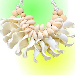 May include: A necklace featuring white petal-shaped elements, light tan seashells, and natural wooden beads strung on white cord. The text kraftykorner is displayed at the top. The design consists of layered white floral-like shapes and shells.