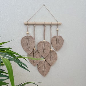 May include: A macrame wall hanging with a wooden dowel and beige leaves. The leaves are made of knotted string and have wooden beads at the top.