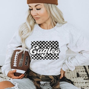 May include: A white sweatshirt with a black and white checkered design and the word "Eagles" in a retro font. The sweatshirt is being held by a person who is also holding a brown football.