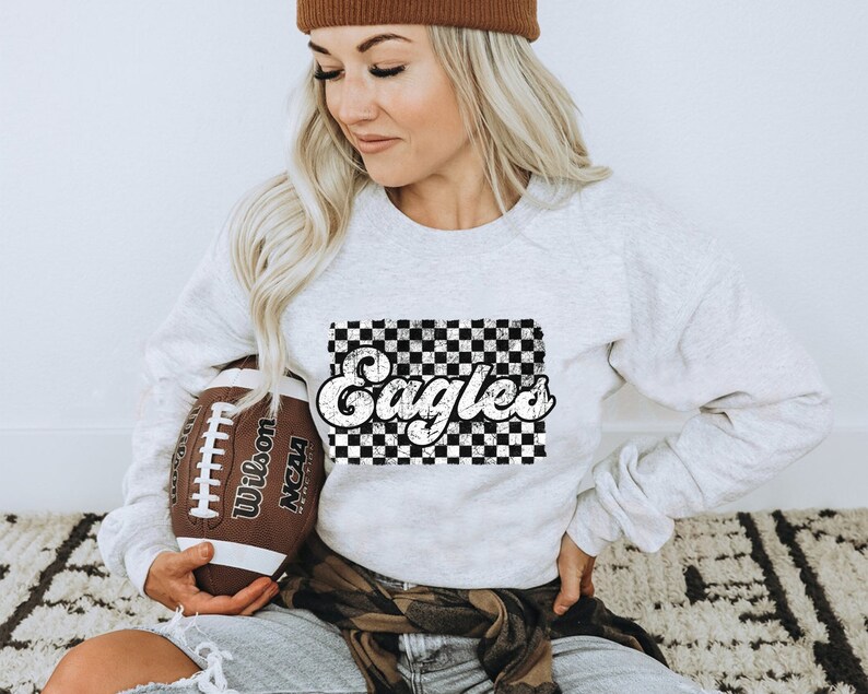 May include: A white sweatshirt with a black and white checkered design and the word "Eagles" in a retro font. The sweatshirt is being held by a person who is also holding a brown football.