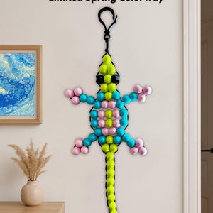 Puede incluir: Una decoraci&oacute;n de lagarto con cuentas, de aproximadamente 112 cm de largo, en una combinaci&oacute;n de colores de primavera limitada. El lagarto presenta un lazo negro para colgar y est&aacute; hecho con cuentas amarillas, rosas, azules y negras. El texto "44" LONG Bead Kit" y "Limited Spring Colorway" son visibles.