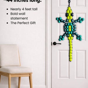 Puede incluir: Un adorno de pared con forma de lagarto hecho con cuentas, de aproximadamente 112 cm de largo, en tonos verdes, turquesas y amarillos, cuelga de una puerta blanca. El texto de la imagen dice "Yes... It's really ~44 inches long."