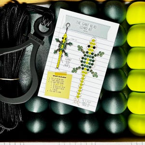 Puede incluir: Un kit de manualidades para crear un lagarto de cuentas, con un diagrama y materiales incluidos. El kit incluye cuerda de nailon, un cierre de colgante, cuentas de poni y otros suministros. El texto en la tarjeta dice "THE GIANT BEAD LIZARD KIT (GREEN)".