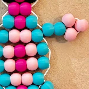 Puede incluir: Una obra de arte con cuentas en forma de &aacute;rbol, con cuentas rosas, turquesas y negras. Las cuentas est&aacute;n ensartadas con un cord&oacute;n blanco, creando un objeto decorativo colorido.