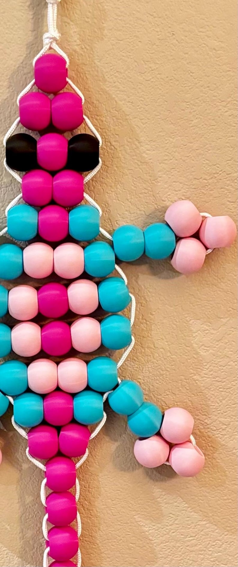 Puede incluir: Una obra de arte con cuentas en forma de &aacute;rbol, con cuentas rosas, turquesas y negras. Las cuentas est&aacute;n ensartadas con un cord&oacute;n blanco, creando un objeto decorativo colorido.