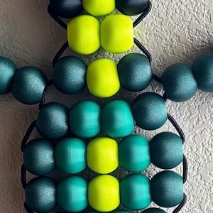 Puede incluir: Un adorno en forma de tortuga hecho de cuentas. La tortuga presenta un cuerpo de cuentas verdes y amarillas, unidas por un cord&oacute;n negro. Las cuentas est&aacute;n dispuestas para formar la cabeza, el cuerpo y las patas de la tortuga. El fondo es una superficie texturizada blanquecina.