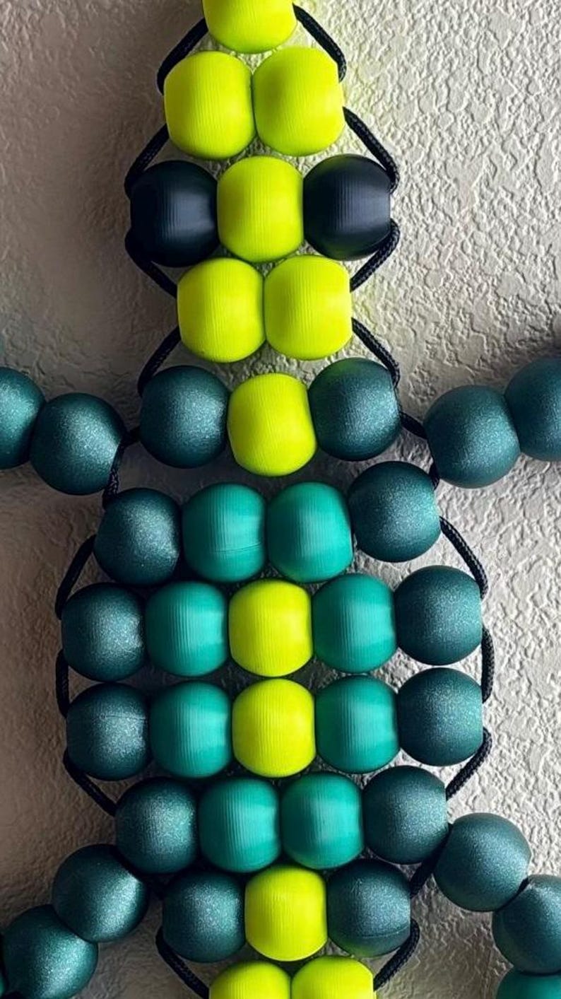 Puede incluir: Un adorno en forma de tortuga hecho de cuentas. La tortuga presenta un cuerpo de cuentas verdes y amarillas, unidas por un cord&oacute;n negro. Las cuentas est&aacute;n dispuestas para formar la cabeza, el cuerpo y las patas de la tortuga. El fondo es una superficie texturizada blanquecina.