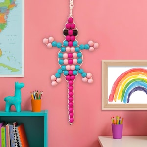Puede incluir: Un lagarto de cuentas para colgar en la pared en tonos rosa, azul y negro. El lagarto est&aacute; suspendido de un lazo blanco. El fondo es una pared rosa con un mapa, una estanter&iacute;a turquesa y un dibujo de arco&iacute;ris enmarcado.