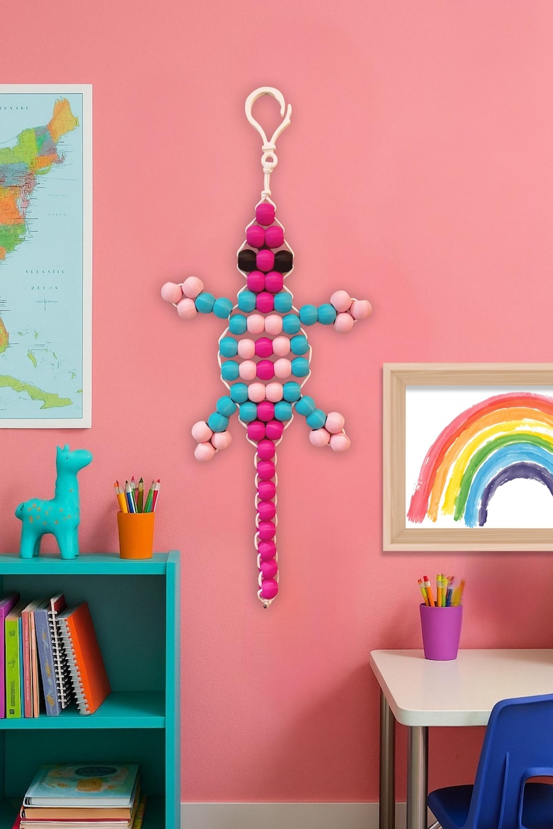 Puede incluir: Un lagarto de cuentas para colgar en la pared en tonos rosa, azul y negro. El lagarto est&aacute; suspendido de un lazo blanco. El fondo es una pared rosa con un mapa, una estanter&iacute;a turquesa y un dibujo de arco&iacute;ris enmarcado.