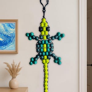 Puede incluir: Un lagarto de cuentas para colgar en la pared en tonos verdes, amarillos y negros. El lagarto est&aacute; suspendido por un lazo negro. El lagarto de cuentas est&aacute; colgado en una pared blanca.