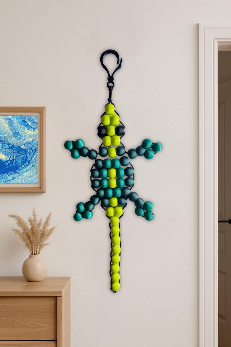 Puede incluir: Un lagarto de cuentas para colgar en la pared en tonos verdes, amarillos y negros. El lagarto est&aacute; suspendido por un lazo negro. El lagarto de cuentas est&aacute; colgado en una pared blanca.