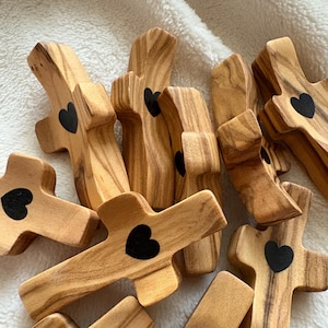May include: A group of ten small wooden crosses with a black heart design. The crosses are made of olive wood and have a natural wood finish.