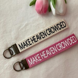 May include: Two keychains with the phrase "MAKE HEAVEN CROWDED" embroidered in black. One keychain is off-white, the other is pink. Both have a bronze-colored metal ring and a matching accent piece. Pink and white tulips are in the background.