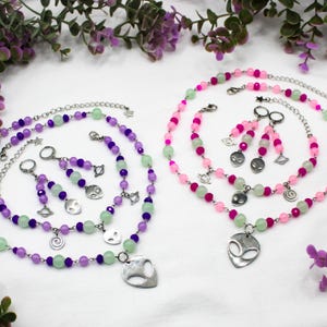 May include: Two sets of beaded jewellery, each with a necklace and earrings. One set is purple and green, the other is pink and green. Each set includes a necklace with an alien pendant, earrings, and a bracelet. The jewellery is on a white surface.