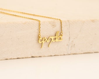 May include: A gold-colored necklace with a delicate chain. The pendant features a stylized script, possibly a name or word. The necklace is displayed on a neutral-toned surface, highlighting its elegant design. The chain is thin and the pendant is in a cursive font.