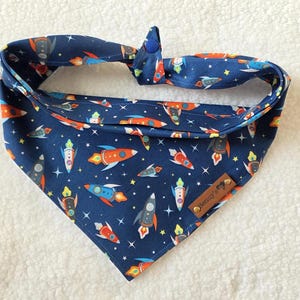 May include: A blue bandana with a pattern of colorful rockets and stars. The bandana has a brown leather tag with the word "Rocket" on it.