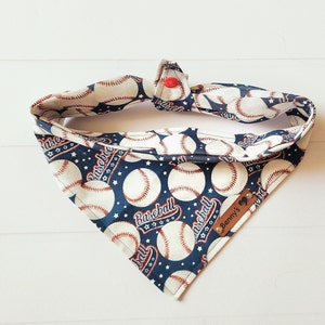 May include: A blue and white bandana with a baseball theme. The bandana features white baseballs with red stitching and the word "Baseball" printed in red. The bandana has a brown leather tag with the name "Benny's" printed on it.