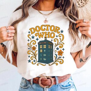 A woman wearing a white t-shirt with a graphic design featuring a blue telephone booth and decorative elements.