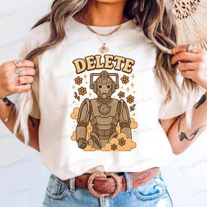 A person wearing a white t-shirt with a graphic design of a robot character and the word "DELETE" printed on it.