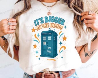 May include: Cream-colored t-shirt featuring a blue police box graphic with the text "IT'S BIGGER ON THE INSIDE" in blue and orange. The design includes stars and the words "BAD WOLF".