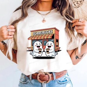 May include: Cream-colored t-shirt featuring a Halloween-themed graphic. The design includes the text "SCAREBUCKS" above two smiling ghost characters holding coffee cups in front of a coffee shop.