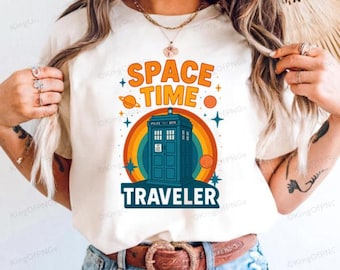 May include: Cream-colored t-shirt featuring a colorful graphic. The design includes the words "SPACE TIME TRAVELER" above an illustration of a blue police box, a rainbow, planets, and stars. The shirt is made of a soft material.