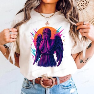 A woman wearing a white t-shirt with a graphic design of a purple angel with wings and a face covering her mouth.