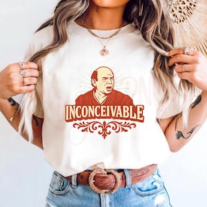 May include: Cream-coloured t-shirt with a red and brown graphic of a man and the word "INCONCEIVABLE" in a decorative font. The shirt has short sleeves and a crew neck.