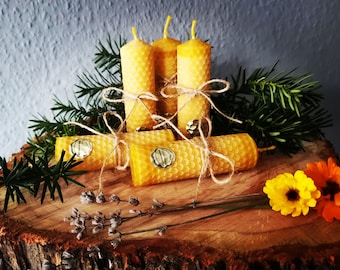 May include: A collection of yellow beeswax candles and rolled candles tied with twine, arranged on a wooden surface with greenery and flowers. The candles have a honeycomb texture and are presented in a rustic, natural style.
